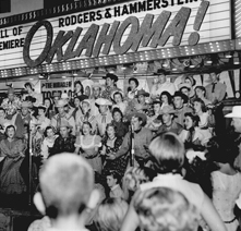 A chorale signing under a movie theater marquee advertising Oklahoma! 