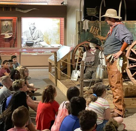 Living history actor dressed as a cowboy speaks to a group of schoolchildren seated in front of him.