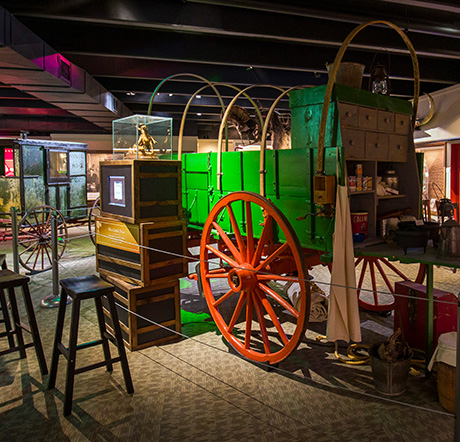 A buckboard-style chuck wagon with wooden bows. The chuck box at the front of the wagon is open, revealing canned goods and equipment. Crates are stacked nearby as part of an exhibit.