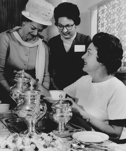 Three women in dresses pour tea from a silver service.