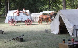 Two men ride in a buckboard wagon through an encampment at Fort Towson.
