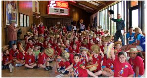 Children wearing cowboy hats salute the camera. They are seated in front of the Will Rogers Theatre.