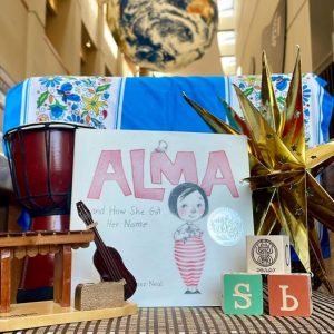 A photo with textiles, drums, instruments, wooden blocks, and a luminaria all surround a book, “Alma: and How She Got Her Name.”
