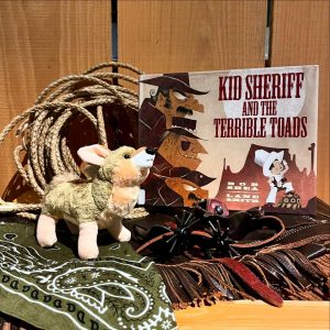 The book 'Kid Sheriff and the Terrible Toads' depicts Western bandits and a child sheriff. A lasso, plush wolf, spurs, chaps, and a bandana surround the book.