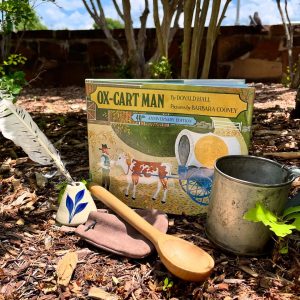 The book 'Ox-Cart Man' depicts a man leading an ox-drawn wagon. An ink quill, leather sack, wooden spoon and metal cup are placed around the book.