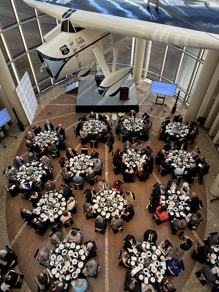 Guests seated at tables for the OHS Awards Banquet, seen from above. The replica of Wiley Post's airplane the "Winnie Mae" is seen suspended overhead.