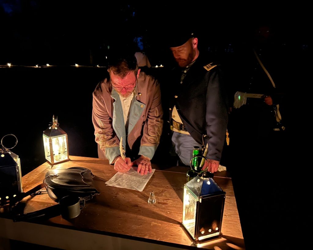 Two reenactors at a lantern-lit table. One is signing a document with an ink quill.