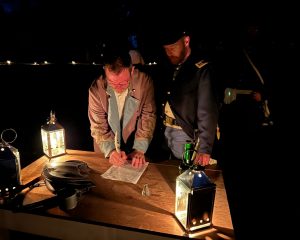 Two reenactors at a lantern-lit table. One is signing a document with an ink quill.