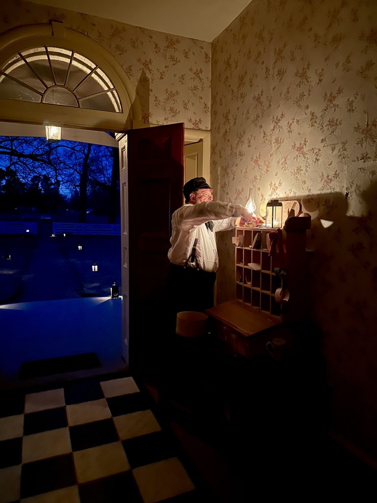 A man lights a lantern at the entrance to Hunter's Home. A path of lights can be seen leading to the entrance lobby where he stands.