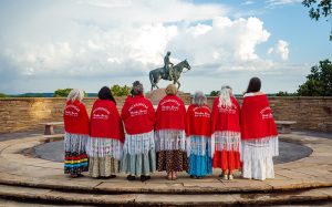 A group of women in red shawls face the Will Rogers tomb at a wreath laying ceremony.