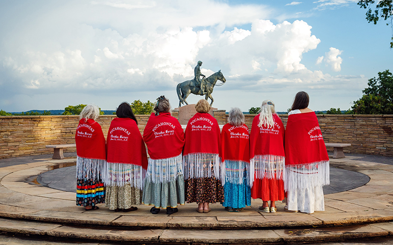 A group of women in red shawls face the Will Rogers tomb at a wreath laying ceremony.