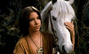 A boy dressed in an ancient garment wears an amulet as he stands by and holds the head of a white horse.