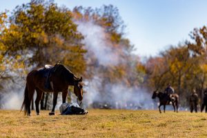 A reenactor lays at the foot of his horse while a reenacted battle occurs in the distance.