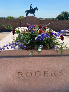 A commemorative wreath with flowers and greenery rests on Will Rogers tomb. The statue of Will Rogers on horseback is seen in the background.
