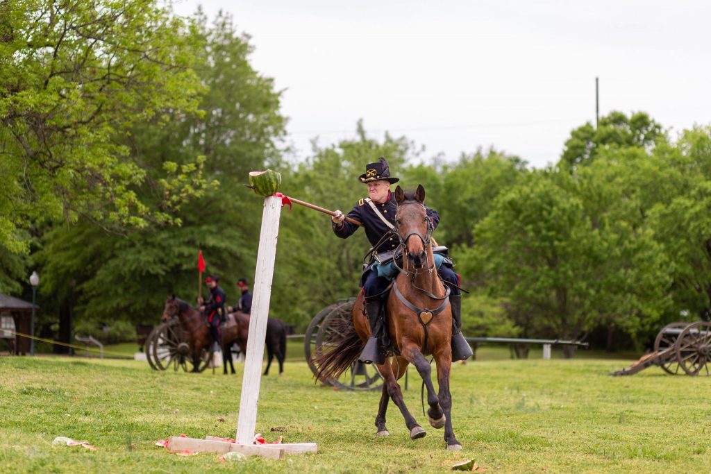 Cody Faber, riding horseback, swipes his sword at a watermelon perched on a post. He is dressed in a Union Civil War uniform.