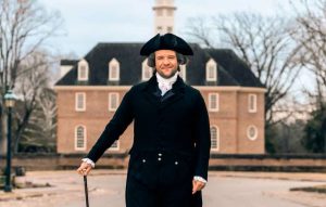 Bryan Austin dressed in Colonial Clothing stands in front of a brick building at Colonial Williamsburg. The living history actor is portraying American founding father James Madison.