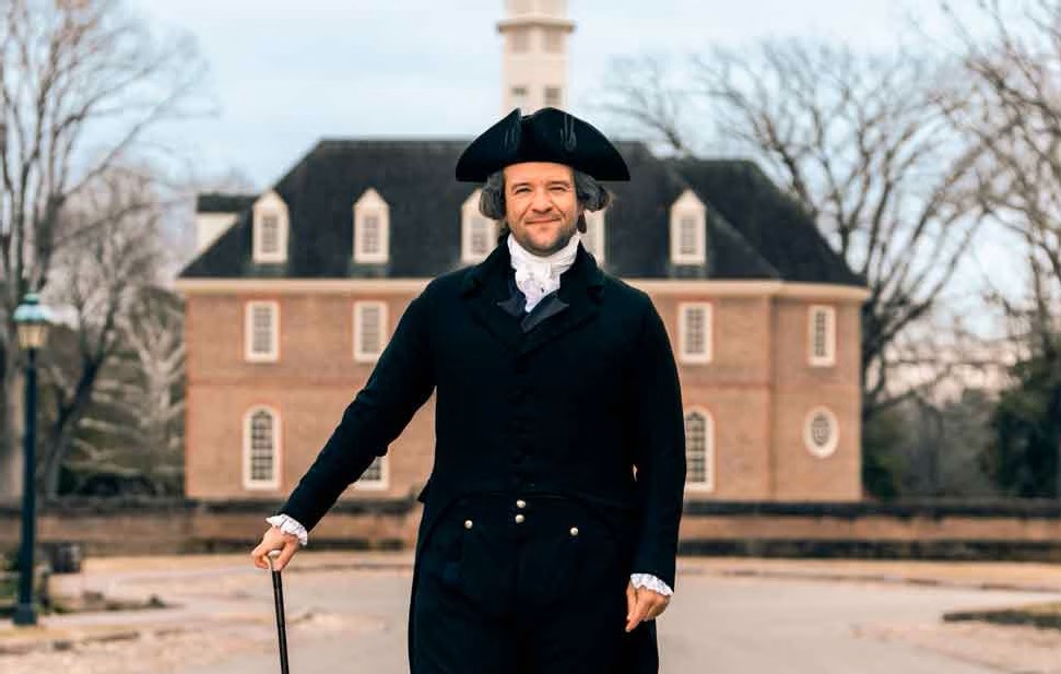 Bryan Austin dressed in Colonial Clothing stands in front of a brick building at Colonial Williamsburg. The living history actor is portraying American founding father James Madison.