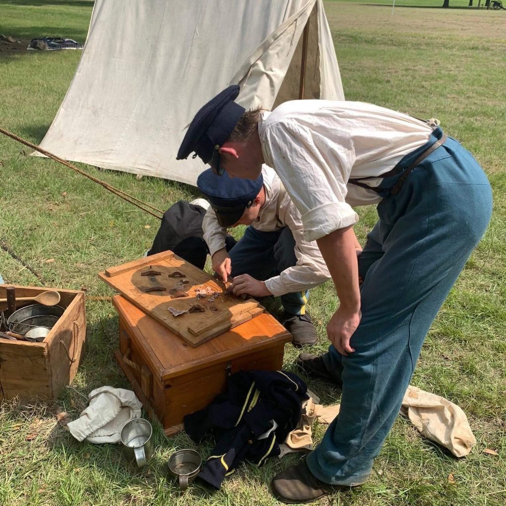 Two soldiers dressed in woolen uniforms work at handcrafting tools.