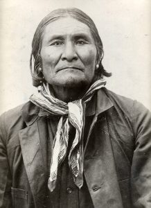 Chiricahua Apache leader Geronimo, looking directly at the camera wearing a coat and scarf around his neck. His hair is shoulder length and he has two earrings in each ear.