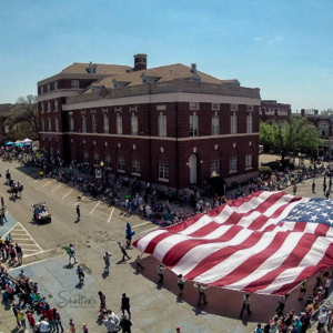 A birdseye view of a large American flag being carried on a Guthrie parade route. The flag stretches from curb to curb.