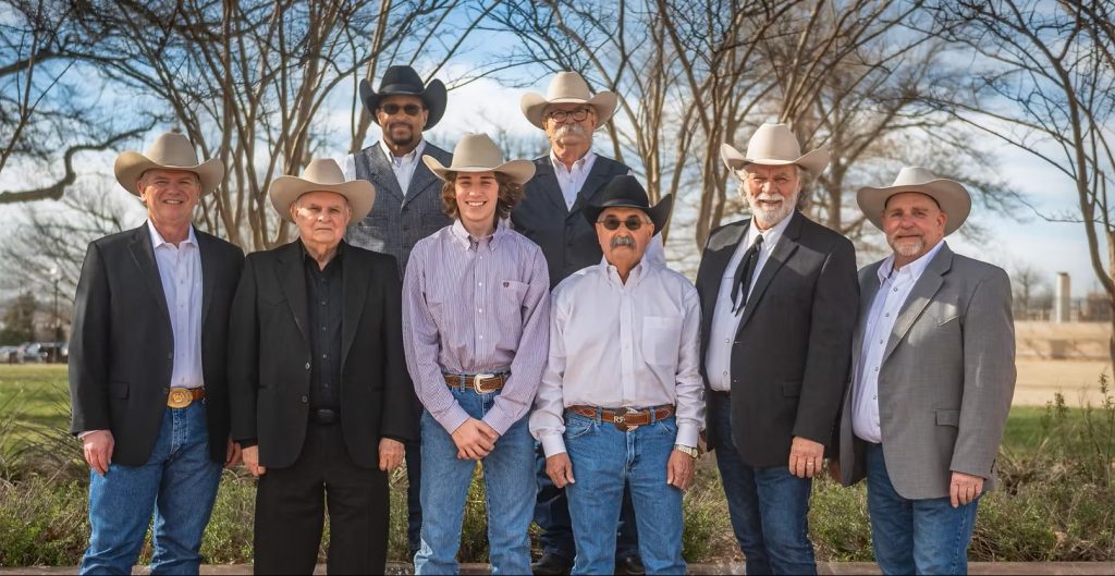 Eight men dressed in Western suits and cowboy hats, standing in two rows.