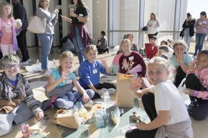 Youngsters seated eating lunch from brown paper bags, waving at the camera.