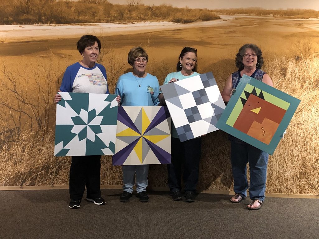 Four women standing together each holding a small 2 foot square barn quilt of their own colorful design.