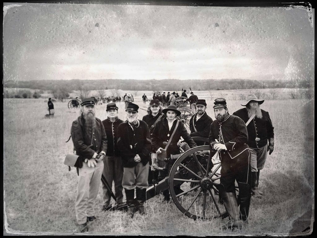 Men gather at a cannon wearing Union Civil War uniforms. Many of the men have beard and all are wearing hats.