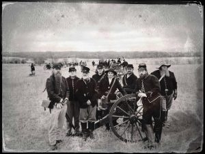 Men gather at a cannon wearing Union Civil War uniforms. Many of the men have beard and all are wearing hats.