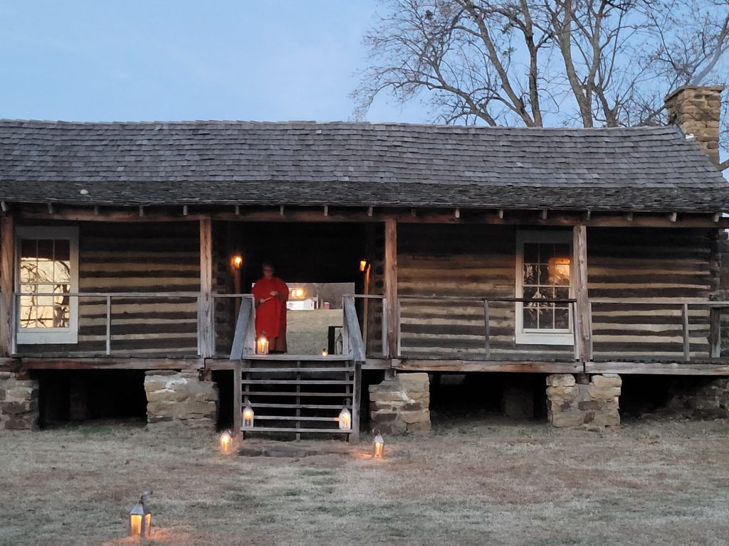 A woman on the porch of a log cabin building lights lanterns on the stairwell leading to the entrance.