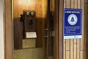 A wooden phone booth with and old fashioned Bell Telephone sign hanging beside it.