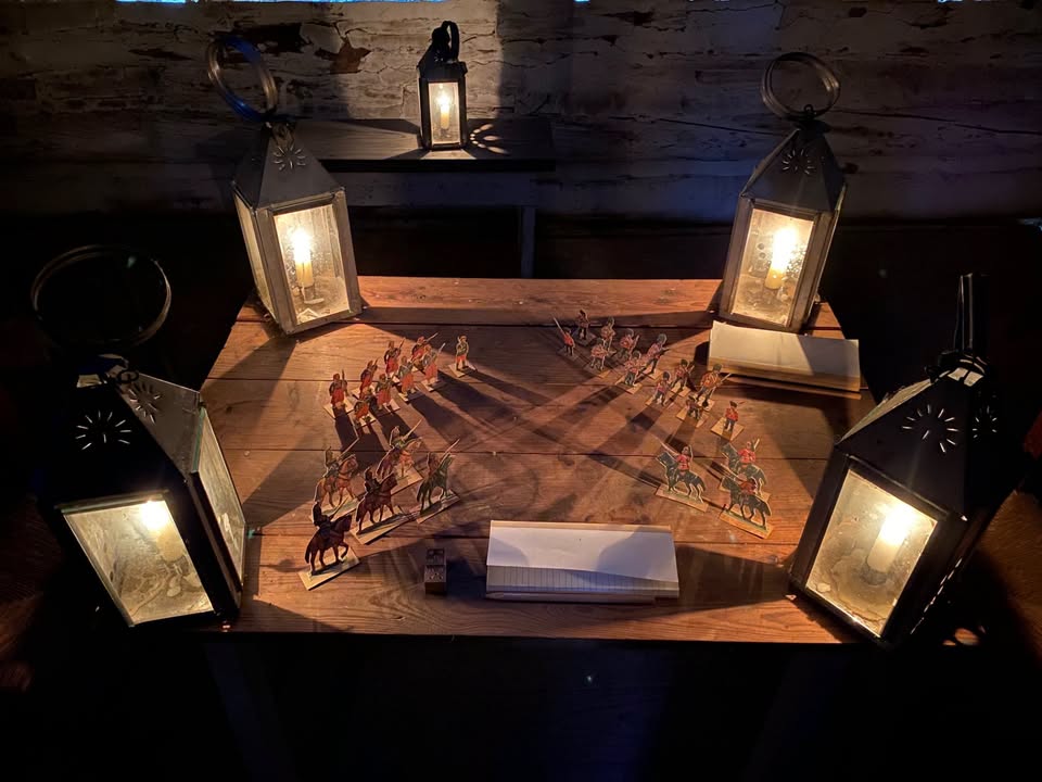 Paper toy soldiers arrayed on a table lit by lanterns with the toy soldiers shadows forming a pattern.