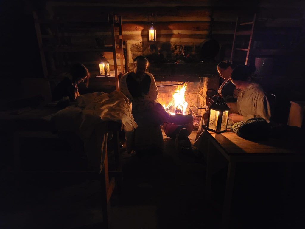 Women gathered at the hearth, sewing by candlelight, with lanterns illuminating the room.