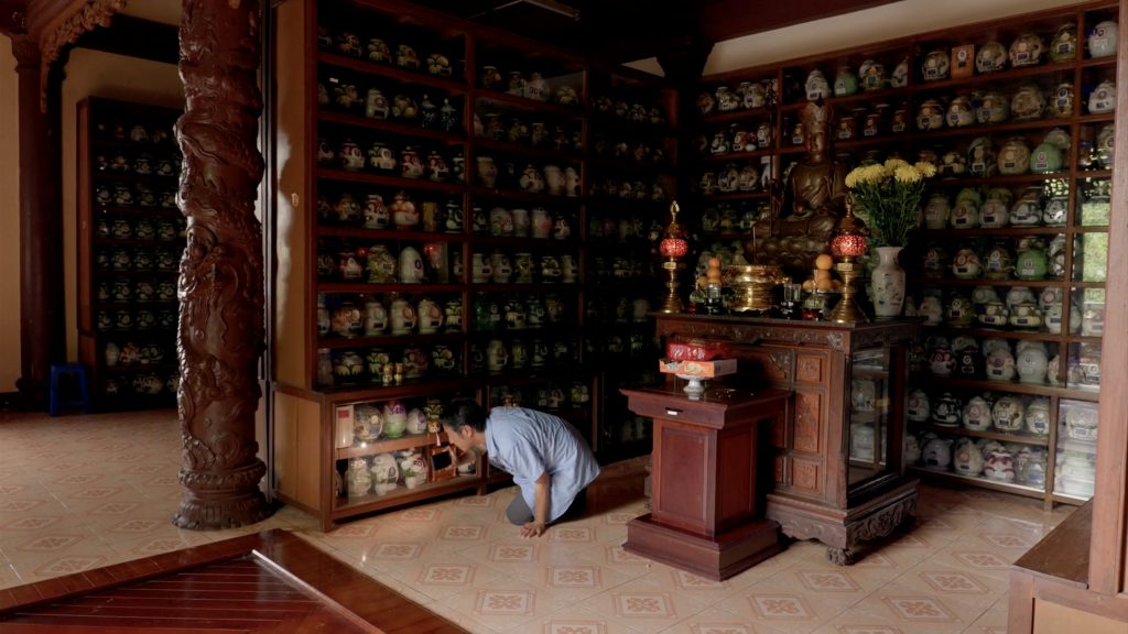 A man, on all fours, holds a cell phone flashlight in his right hand searching multiple ceramic vessels on multilayered shelves.