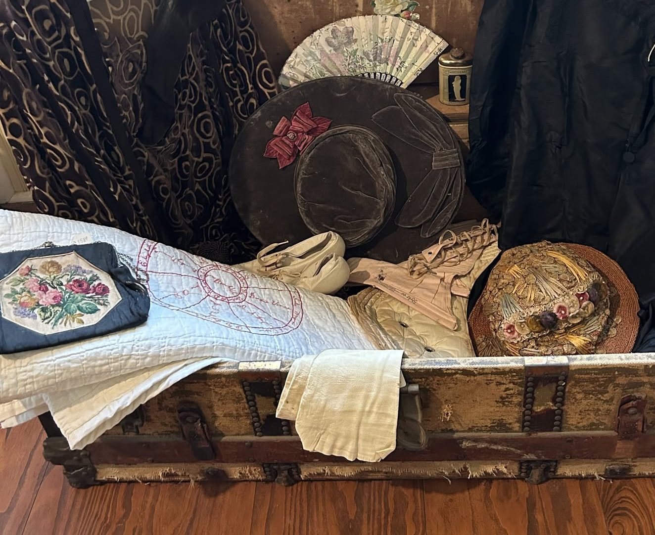 A large vintage traveling trunk with a quilt, hat, purse, gloves, and clothing.