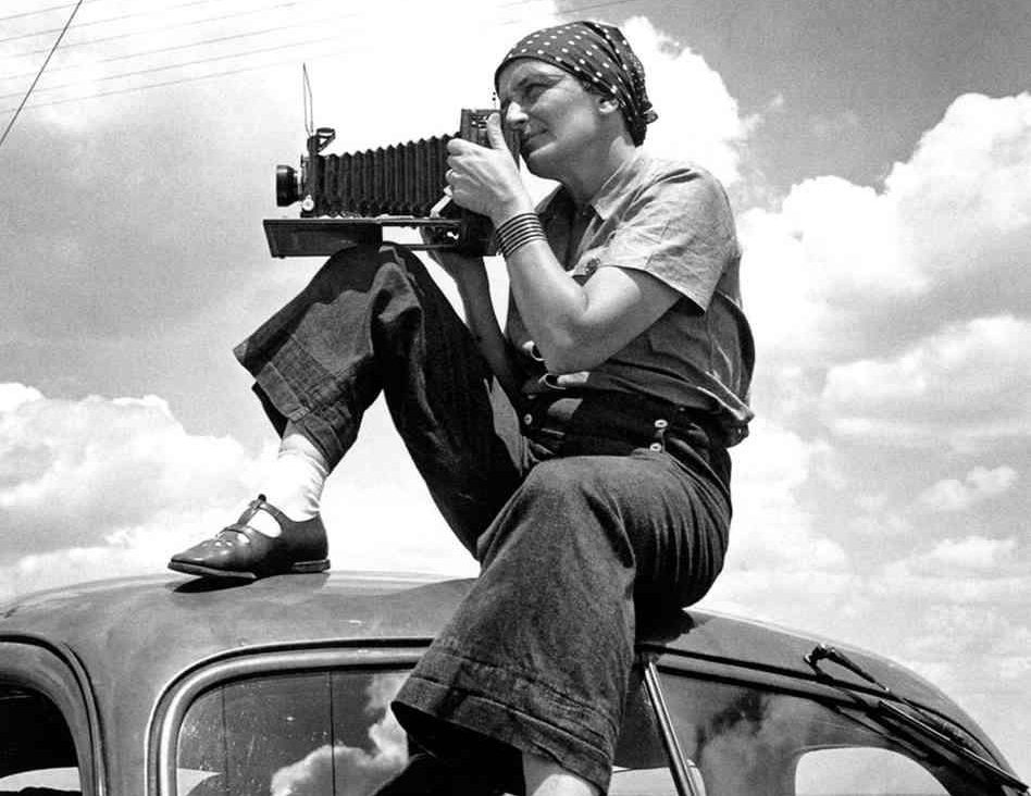Dorothea Lange sits on the roof of a car taking a photograph.