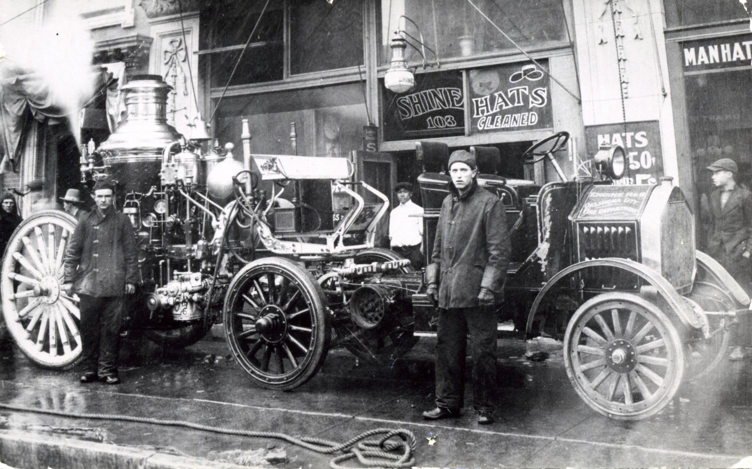 Two men stand in front of an elaborate fire engine with shops and passersby visible in the background.