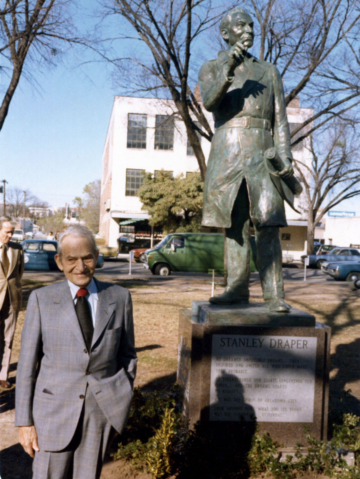 A smiling Draper stands in a suit and tie next to a bronze state of him. The statue holds rolled papers in one hand and points forward with the other. A building, cars, and a man stand in the background.