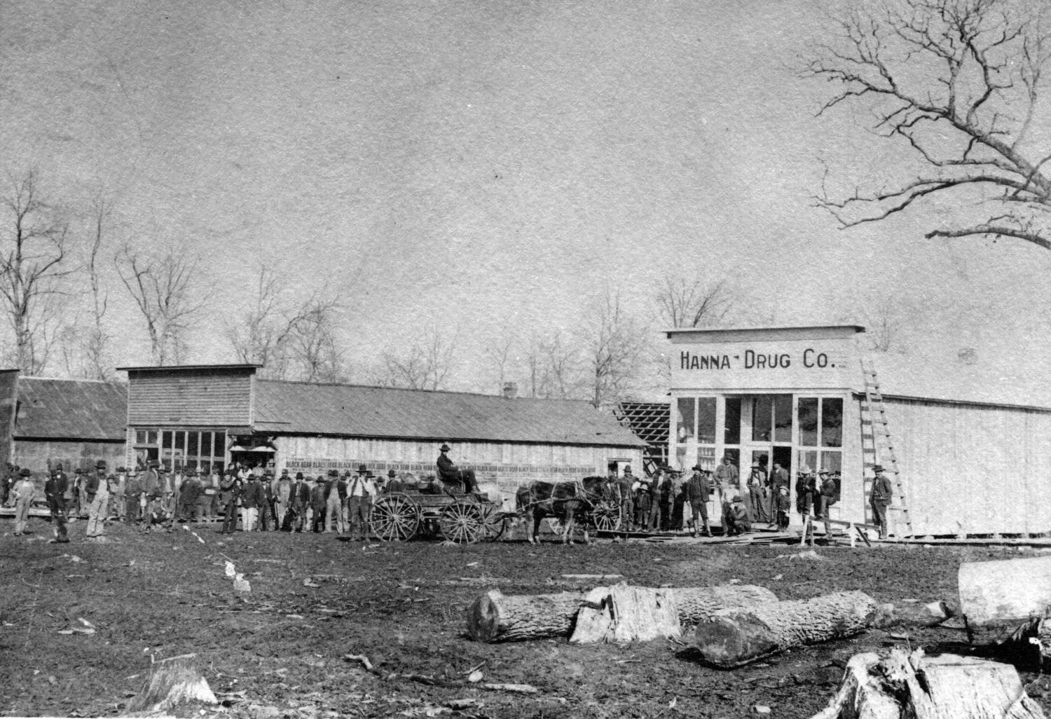 A line of people and a man on a horse-drawn cart stand in front of the Hanna Drug Co., a small wooden structure. Large sections of tree trunks lay dirt in the foreground. Two other structures stand next to the drug store.