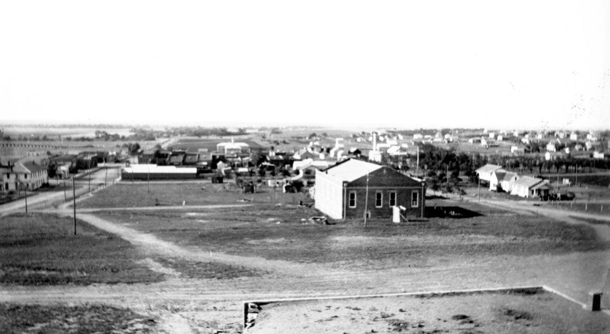 An overhead view of the town with a larger structure in the foreground on a grassy lot, with dirt roads and other smaller buildings in the distance.