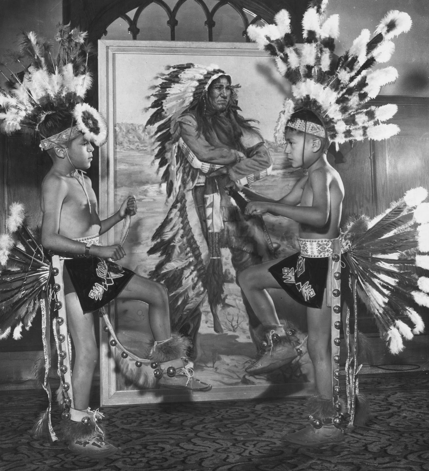Two young boys in traditional Native American dance regalia face each other in front of a painting of a Native man. The boys wear feathered headdresses, a feathered back bustle, and bells.