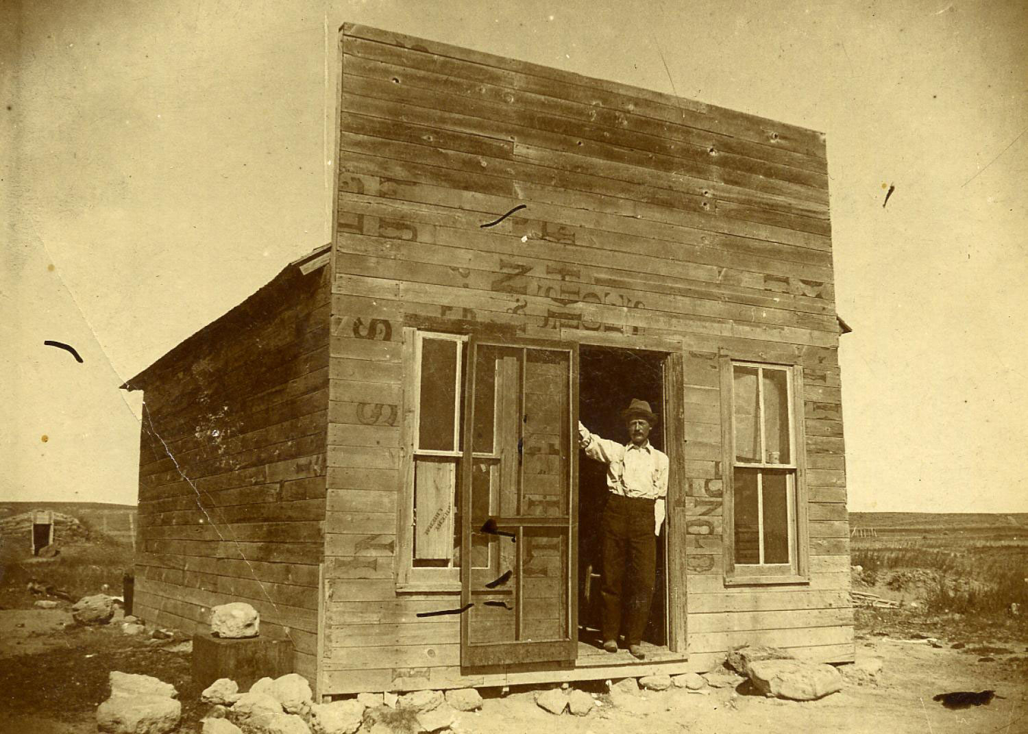 A man stands in the doorway of a very small wooden structure with two windows. The surrounding landscape includes large rocks, grass, and a dugout in the distance.