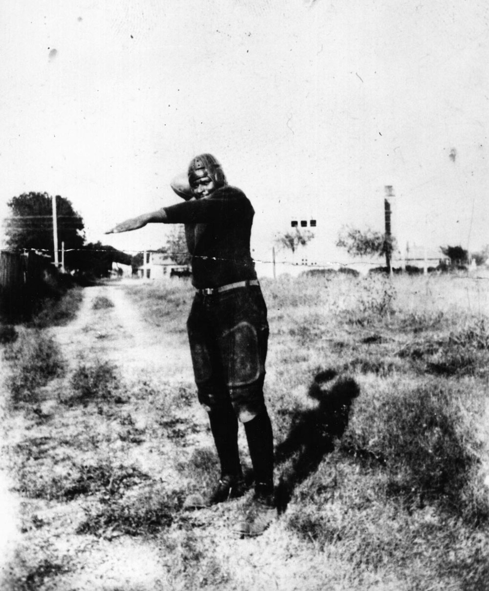 Hall as a young man in a football uniform. He holds a football in one hand, his arm drawn back about to throw. The other arm is stretched out straight in front of him.
