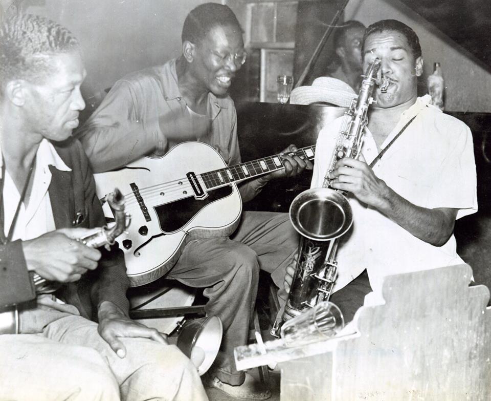 Historic photograph of musician Charlie Christian on the guitar while a man plays the saxapohone and a another man looks on