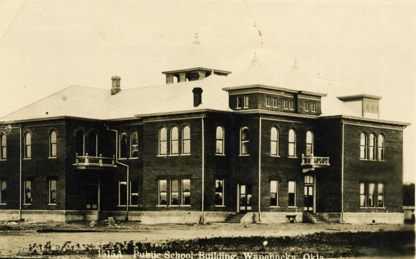 A large two-story brick structure with a tower and arched windows on a dirt lot.