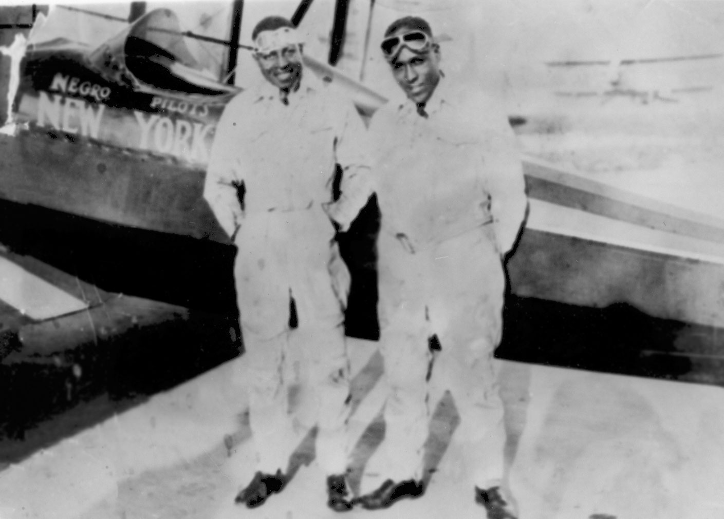Pilots Thomas Allen and James Herman Banning stand in front of an airplane wearing pilots googles and flight suits 