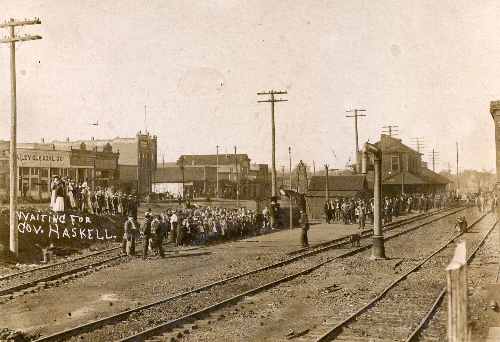 Groups of well-dressed men and women stand along railroad tracks. Behnid them are various commercial buildings and uitility poles. Handwriting on the image reads