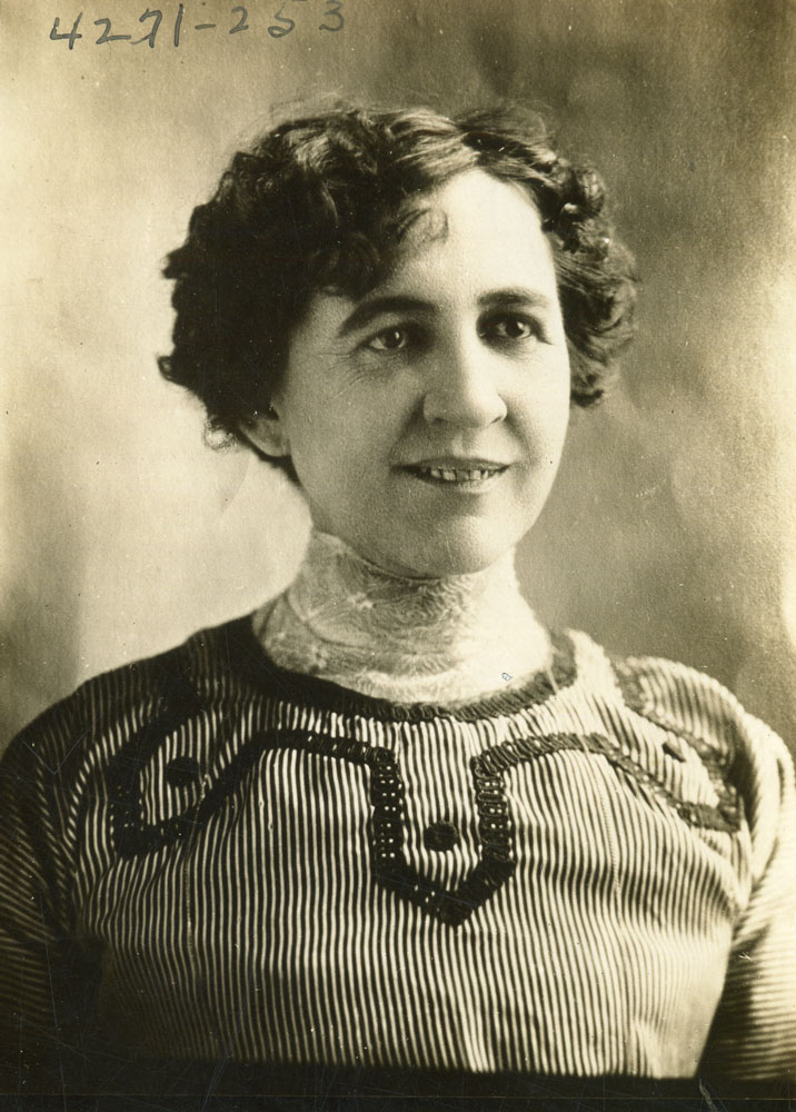 A portrait of Sturgeon with a small smile. She wears a striped dress with embroidered details and a lace collar. Her curly hair is in an updo.