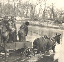 Men in a simple wagon are pulled across a river by two mules