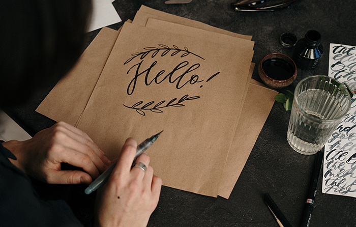 Hand holding a calligraphy pen, and a brown paper with a hand-written Hello! and decorative leaves